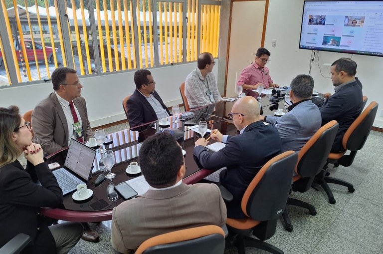 Foto mostra pessoas em reunião com o apoio de um telão