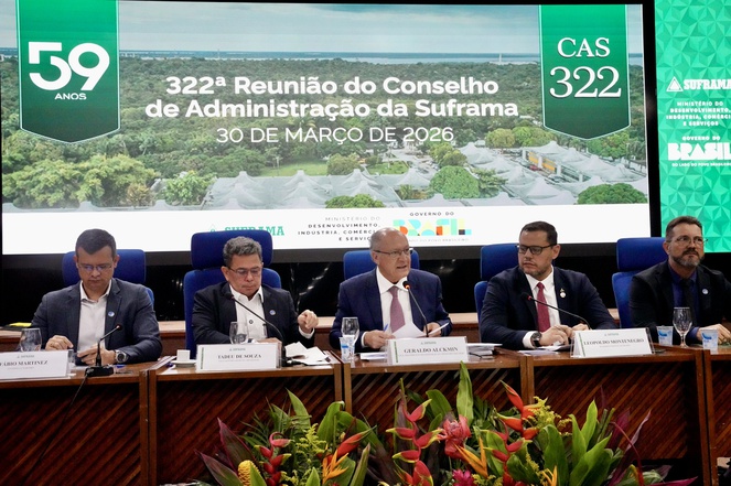 Foto mostra homem de paletó e gravata falando e observado por outras pessoas, com um telão ao fundo