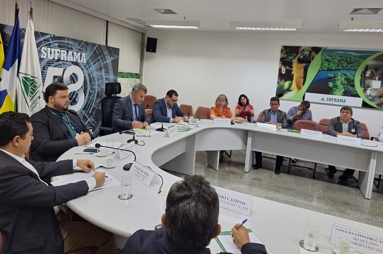 Foto mostra homem de paletó e gravata falando ao microfone durante uma reunião