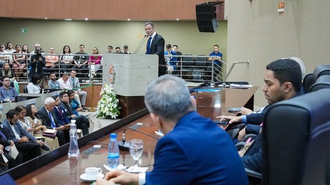 Foto mostra homem de paletó preto flando ao microfone em evento público