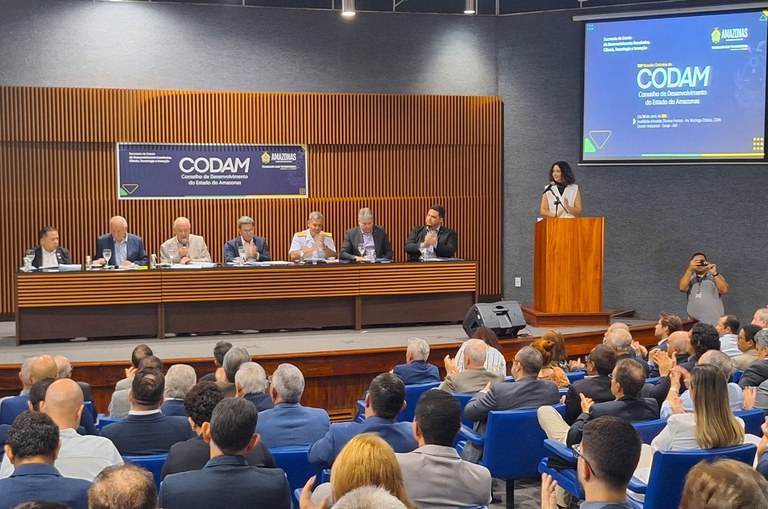 Foto mostra homem de blazer claro falando ao microfone, observado por outras pessoas 