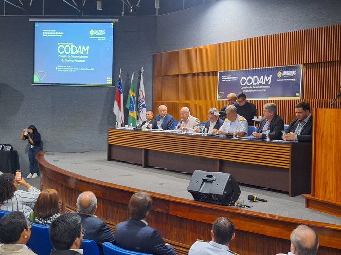 Foto mostra homem de blazer claro falando ao microfone em um evento público