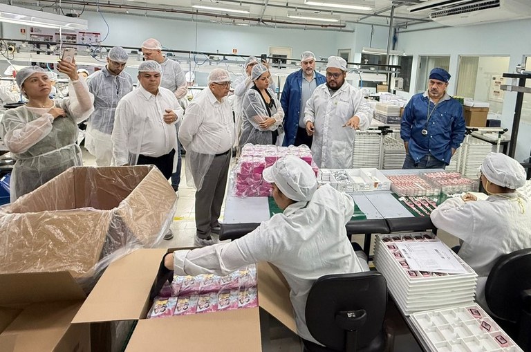 Foto mostra grupo de pessoas com batas brancas e azuis observando produtos em uma fábrica