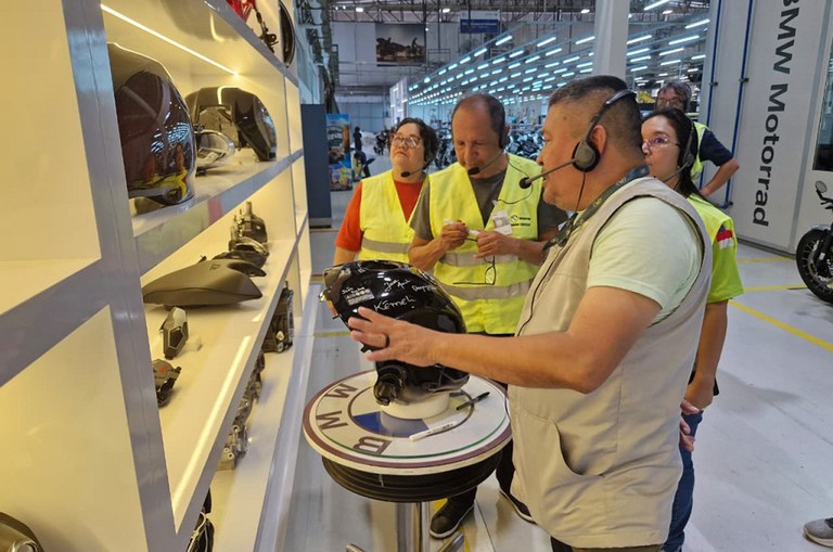 Três pessoas com coletes amarelos observam um homem demonstrando um capacete preto dentro de uma fábrica da BMW Motorrad, com peças expostas em prateleiras e ambiente industrial ao fundo.