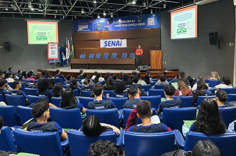 Foto mostra mulher de blusa laranja falando a estudantes em um auditório