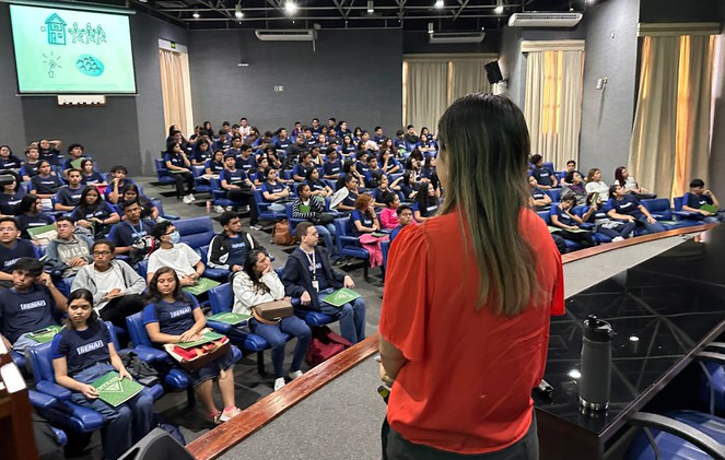 Foto mostra mulher de blusa laranja em pé, observada por grupo de estudantes