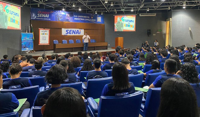 Foto mostra homem em pé falando ao microfone num auditório lotado