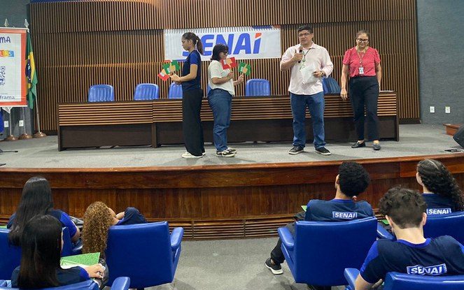 Foto mostra alunos segurando placas e um homem falando ao microfone