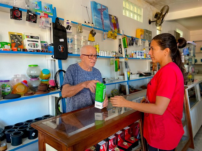 Foto mostra um senhor de óculos oferendo produto a uma cliente vestida de blusa vermelha num estabelecimento comercial