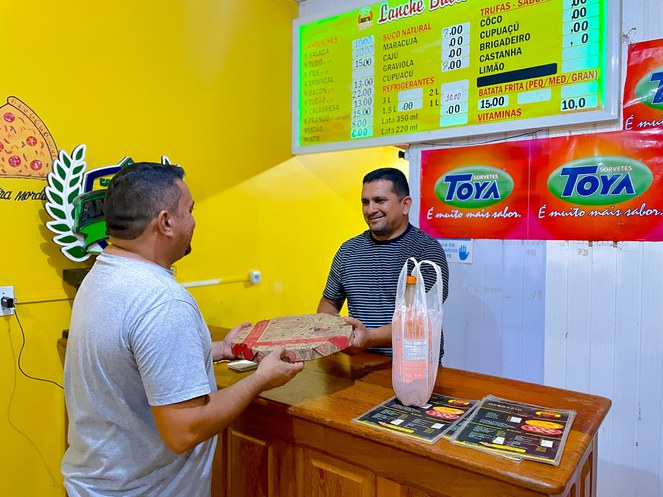 Foto mostra comerciante entregando uma pizza a cliente num estabelecimento comercial