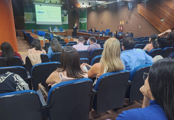 Foto mostra pessoas olhando para apresentação de um homem vestido de blazer e caça escuros