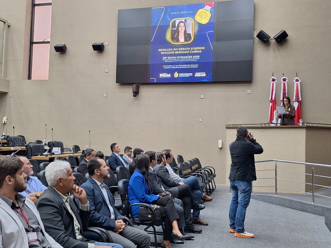 Foto mostra mulher falando numa tribuna observada por grupo de pessoas