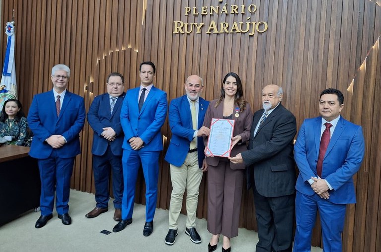 Foto mostra mulher de blazer segurando um documento ao lado de um grupo de homens