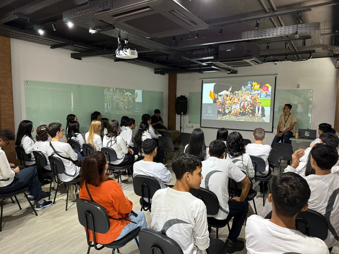 Foto mostra grupo de alunos de blusa branca e um telão ao fundo