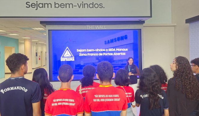 Foto mostra mulher fazendo uma explicação a grupo de estudantes, com um painel ao fundo
