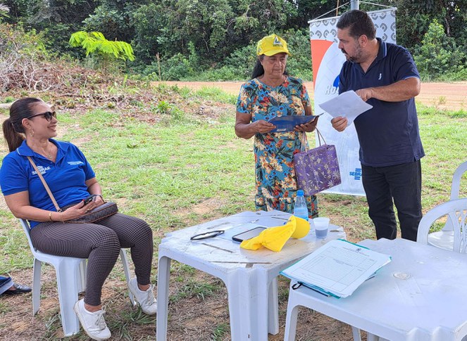 Foto mostra homem de blusa azul segurando um papel e fazendo uma explicação a uma senhora