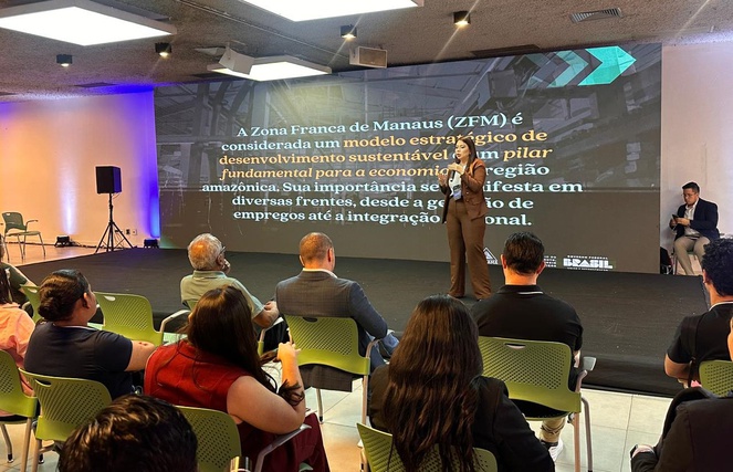 Foto mostra mulher de blazer falando em público com um telão como apoio