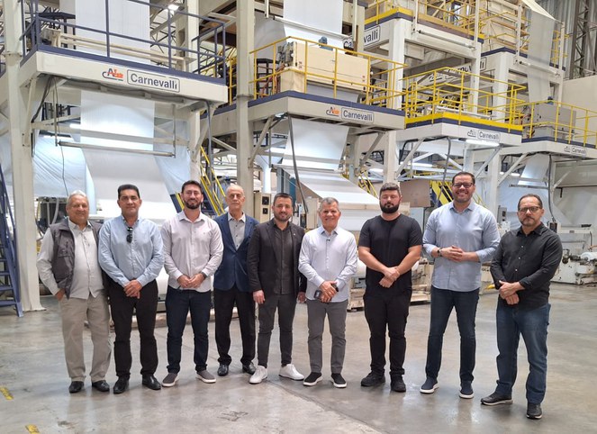 Foto mostra grupo de homens posando para fotografia dentro de uma fábrica com produtos plásticos