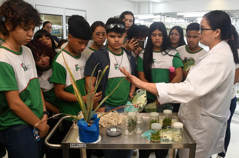 Foto mostra mulher de jaleco branco mostrando planta a estudantes