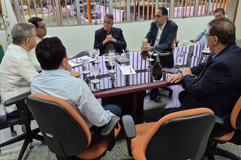 Foto mostra homem de blazer azul e óculos falando a outro homens em uma reunião