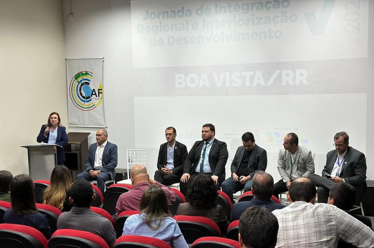 Foto mostra mulher de blazer falando ao microfone em um aditório lotado e com um telão ao fundo