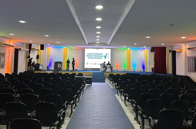 Foto mostra um auditório sendo preparado para um evento, com um grande painel ao fundo