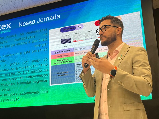 Foto mostra homem falando ao microfone com um painel grande ao fundo