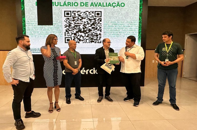 Foto mostra homem falando ao microfone, observado por outro homem com livro na mão