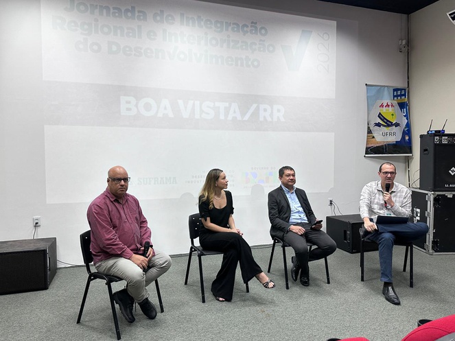 Foto mostra homem sentado falando ao micorfone ao lado de outras pessoas e um telão ao fundo