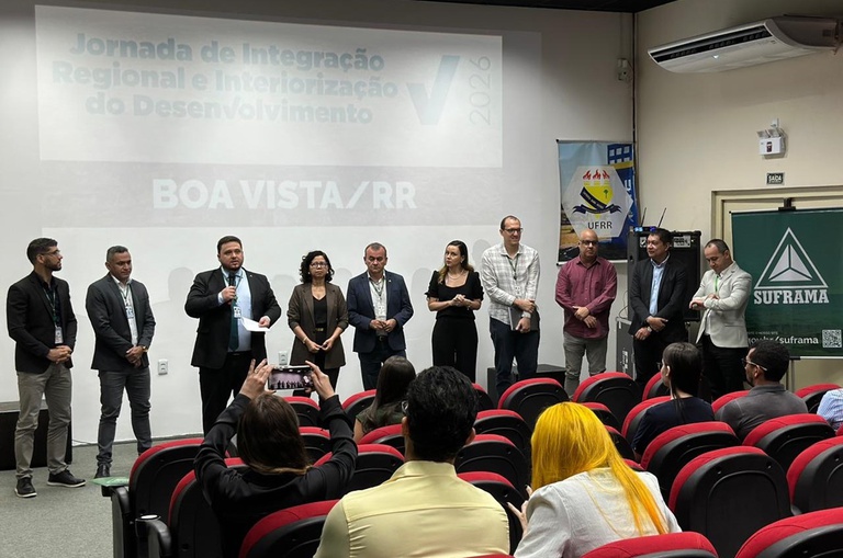 Foto mostra homem de blazer falando ao microfone, observado por grupo de pessoas e um telão ao fundo