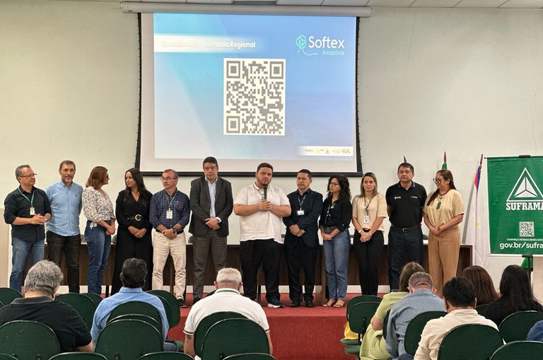 Foto mostra uma perspectiva a partir do fundo de um auditório, onde aparecem as cadeiras das primeiras fileiras e, no primeiro plano, o palco, onde estão doze pessoas de pé sendo que a sétima, da esquerda para a direita, está falando ao microfone. No fundo do palco um telão exibe um QR-Code e, na lateral direta, há um banner com a marca da Suframa.