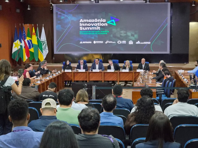 Foto mostra um homem de blazer azul falando ao microfone, com um telão ao fundo e pessoas ao redor