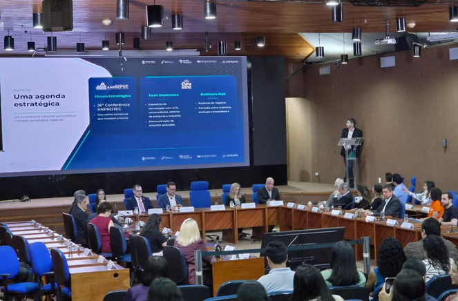 Foto mostra homem de blazer preto em pé e falando ao microfone, com um telão ao como apoio e pessoas ao redor