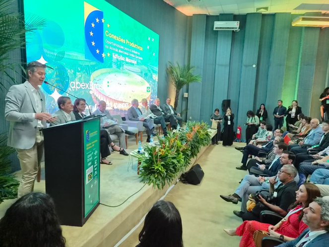 Foto mostra homem em pé falando em evento público, num auditório lotado
