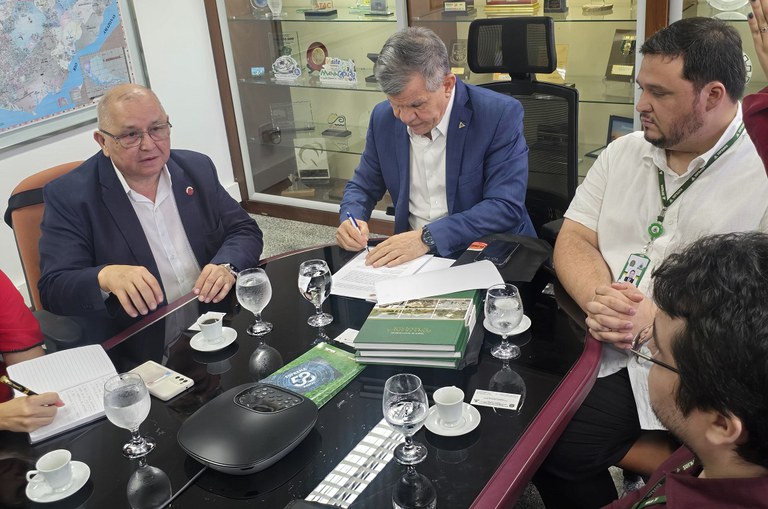 Foto mostra homem assinando documento, observados por outro grupo de homens