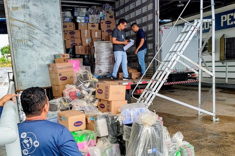 Foto mostra homem olhando para dentro de um caminhão cheio de mercadorias onde outro homem anota numa prancheta as informações sobre os produtos