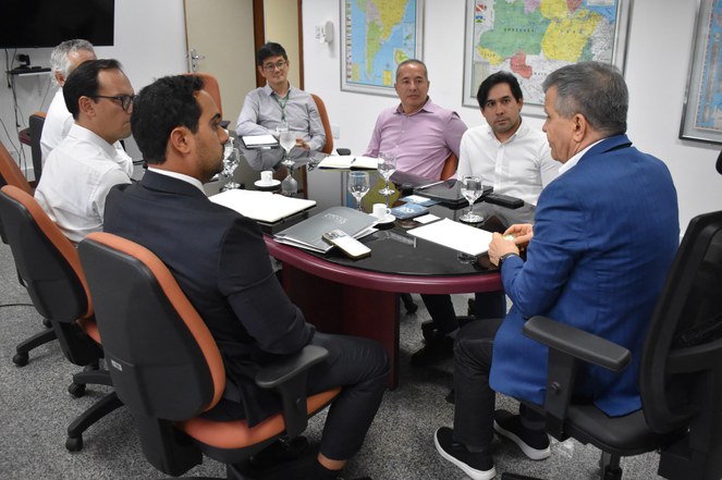 Foto mostra homem de paletó azul escuro em reunião com outros homens, numa sala de reuniões fechada e com mapas geográficos de fundo