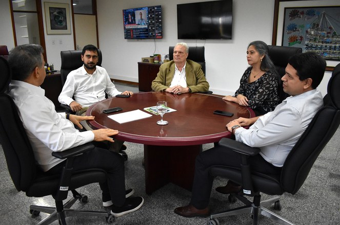 Foto mostra homem de blusa branca e calça preta, conversando com outros três homens e uma mulher durante uma reunião