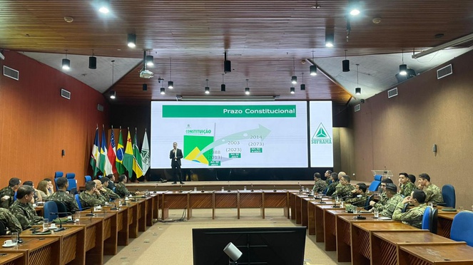 Foto mostra homem em pé fazendo uma palestra a grupo de pessoas numa reunião, com um telão de apoio