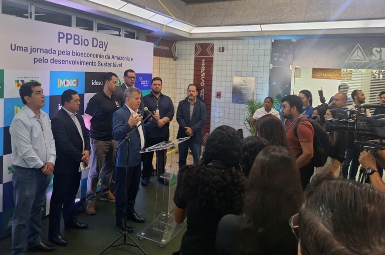 Foto mostra homem de paletó falando ao microfone, observado por por grupo de pessoas