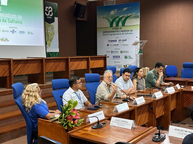 Foto mostra homem falando ao microfone com telão ao fundo e observado por outras pessoas