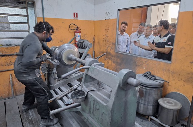 Foto mostra homem mexendo numa máquina, observado por outros homens atrás de uma janela