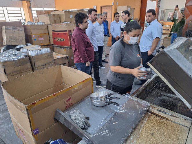 Foto mostra mulher com máscara mexendo em panelas, observa por um grupo de homens