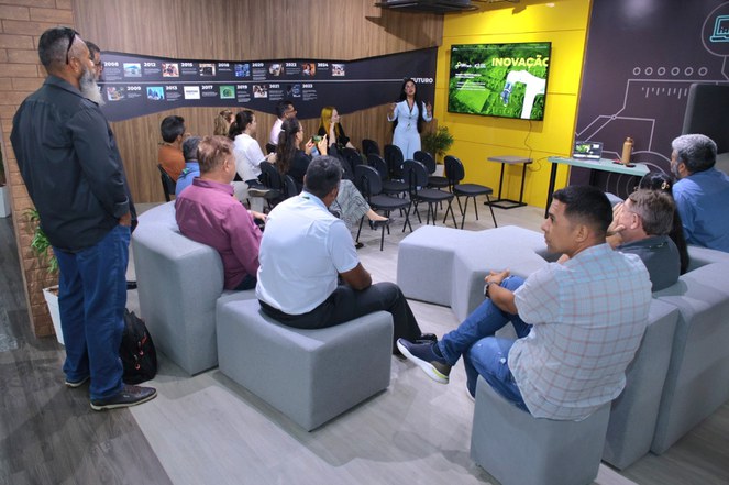 Foto mostra mulher de branco fazendo uma explanação a um grupo de pessoas com um telão como apoio