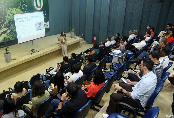 Foto mostra uma mulher de roupa bege fazendo uma palestra em auditório cheio, com o apoio de um telão