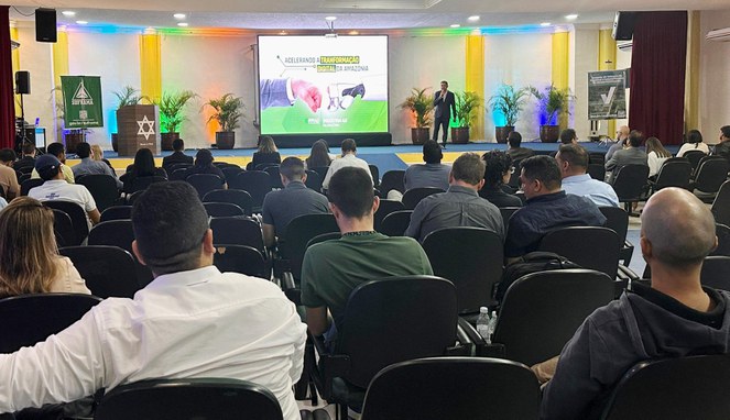 Foto mostra um homem falando com microfone na mão a um grupo de pessoas, com um grande painel ao fundo