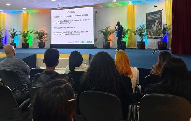 Foto mostra homem com microfone na mão falando a um grupo de pessoas, com um grande painel ao fundo