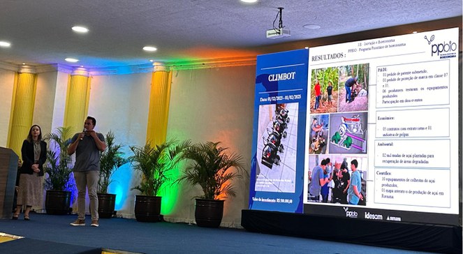 Foto mostra homem falando com microfone na mão e ao lado de uma mulher, a um grupo de pessoas, com um grande painel ao fundo