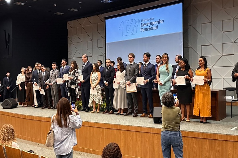 Foto mostra pessoas num palco com certificados nas mãos, sendo fotografadas e com um telão ao fundo