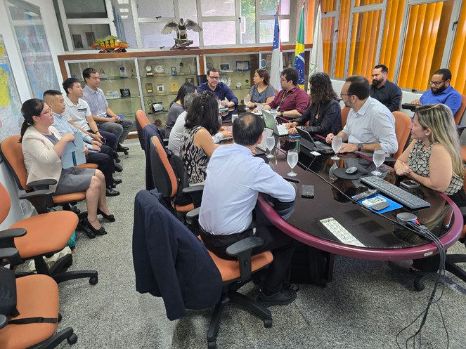 Foto mostra homem de blusa azul, ao centro da mesa falando a grupo de pessoas numa reunião